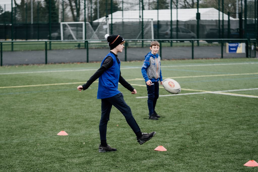 A young boy on a sports field kicking a rugby ball.
